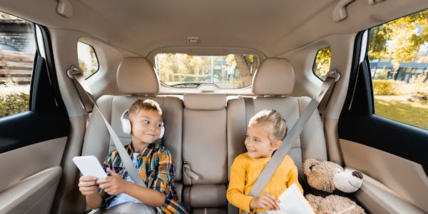 Two children sitting in the back of a car with a wooden aircraft and a teddy bear