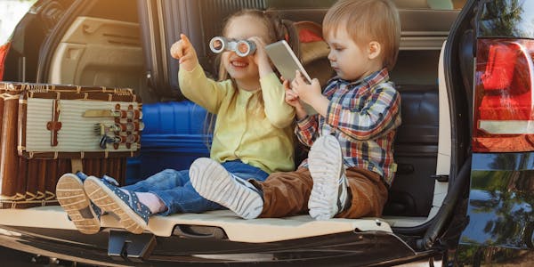 Two kids sitting in the boot of a car, one playing games and one with binoculars