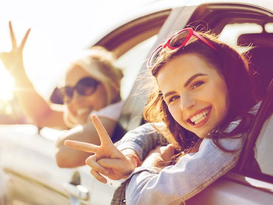 Two girls, giving the peace sign, leaning out of a static car window whilst the sun shines